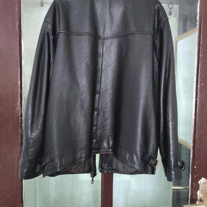 May include: A black leather jacket with a classic design. The jacket features a collar, a zipper closure, and a belt detail at the waist. The jacket is hanging on a door.