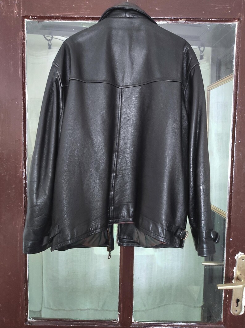 May include: A black leather jacket with a classic design. The jacket features a collar, a zipper closure, and a belt detail at the waist. The jacket is hanging on a door.