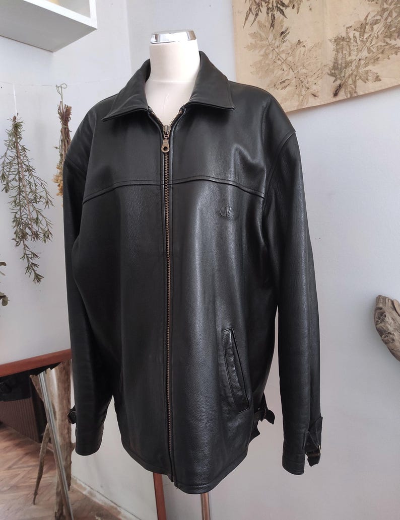 May include: Black leather jacket with a zip-up front, collar, and side pockets. The jacket has a small logo on the chest and adjustable cuffs. The jacket is displayed on a mannequin.