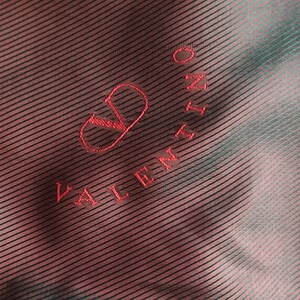 May include: Close-up of a fabric with a diagonal striped pattern in shades of red and teal. The Valentino logo is embroidered in red thread, featuring the brand name and a stylized 'V' symbol.