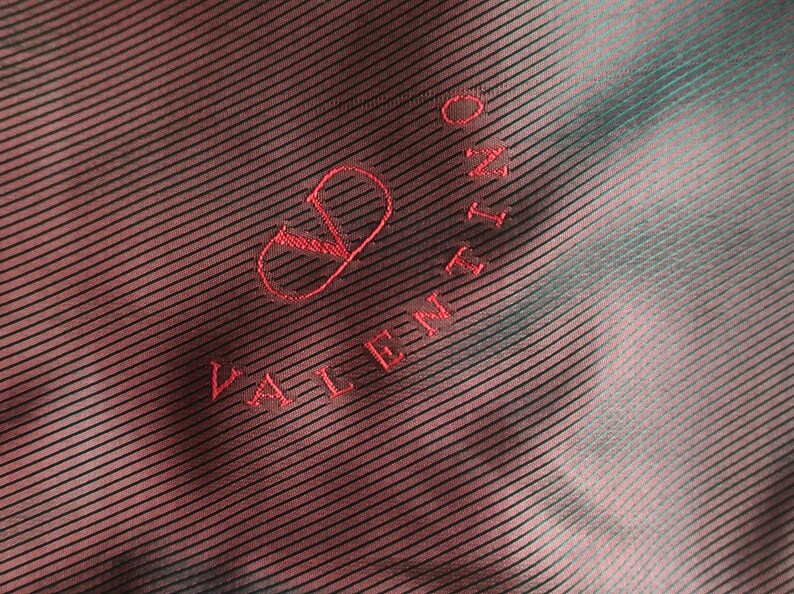 May include: Close-up of a fabric with a diagonal striped pattern in shades of red and teal. The Valentino logo is embroidered in red thread, featuring the brand name and a stylized 'V' symbol.