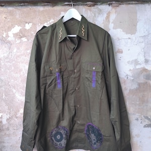 Può includere: Camicia verde oliva con cuciture decorative sul colletto e sulle tasche. Presenta dettagli viola e toppe vicino all'orlo. La camicia è appesa a una gruccia bianca.