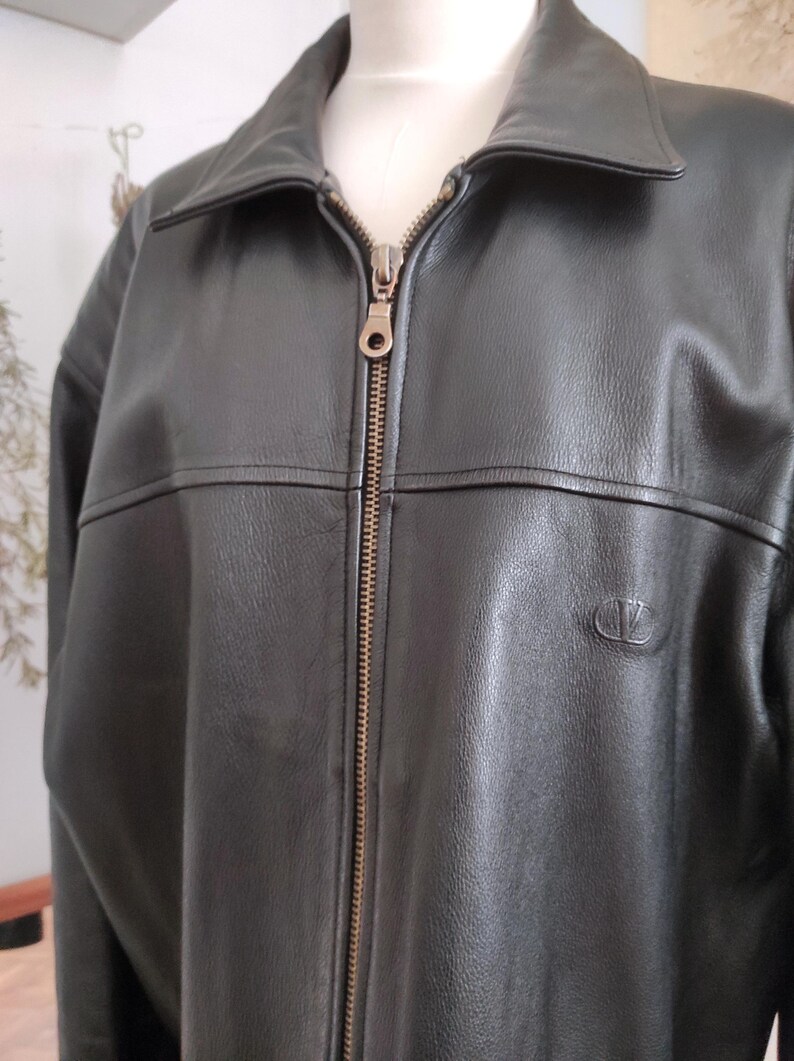 May include: Black leather jacket with a collared neckline and a full-length zipper. The jacket has a small logo on the chest and a classic design. The jacket is displayed on a mannequin.