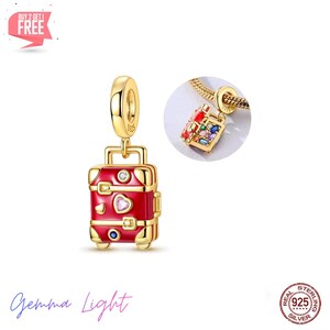 Puede incluir: Un charm dorado con forma de maleta roja con detalles dorados. El charm de la maleta tiene una gema rosa en forma de corazón, una gema azul redonda y una gema transparente redonda. También se muestra un charm más pequeño con gemas de colores.