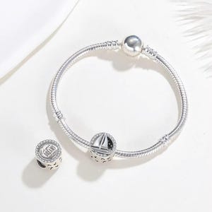 May include: A silver snake chain bracelet featuring a round charm with a sailboat design and a separate round charm engraved with the text Let your DREAMS set sail, both accented with small clear stones along the edges of the charm surfaces.