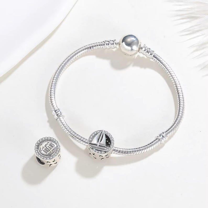 May include: A silver snake chain bracelet featuring a round charm with a sailboat design and a separate round charm engraved with the text Let your DREAMS set sail, both accented with small clear stones along the edges of the charm surfaces.