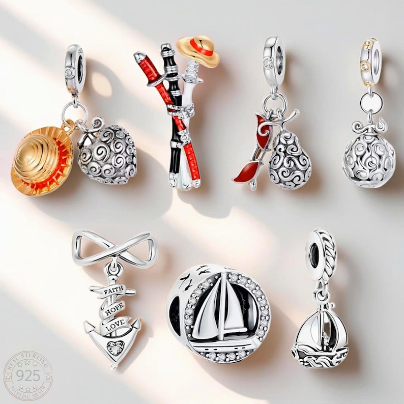 May include: A collection of seven silver charms featuring various designs including a gold-coloured straw hat, a red and black sword, a pear-shaped fruit, an anchor with the words FAITH, HOPE, and LOVE, and two sailboat motifs with clear stones.