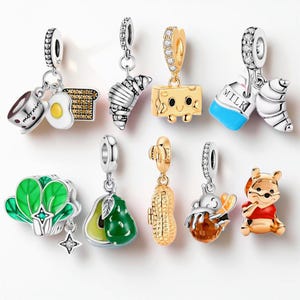 May include: A collection of silver and gold-toned metal charms featuring various food and character designs, including a coffee cup with the word morning, a fried egg, toast, a croissant, a cheese block, a milk carton, bok choy, an avocado, a peanut, a bee, and a bear.