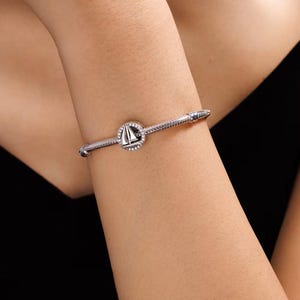 May include: A silver-toned snake chain bracelet featuring a circular charm with a sailboat motif. The charm is accented with small, clear stones set around the perimeter of the circle, and the bracelet has a secure, threaded clasp mechanism.