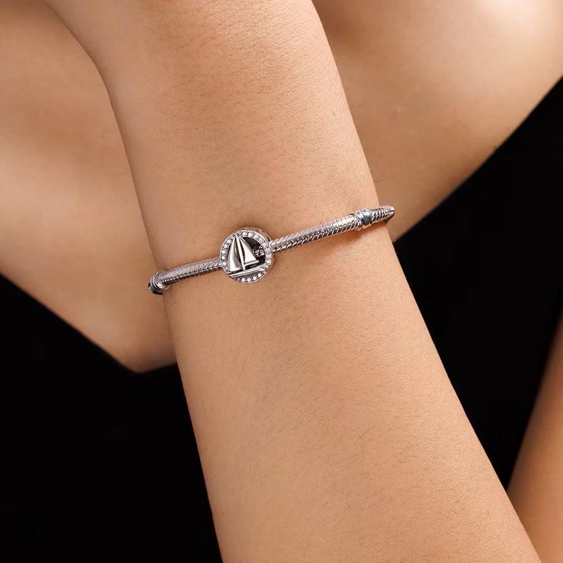 May include: A silver-toned snake chain bracelet featuring a circular charm with a sailboat motif. The charm is accented with small, clear stones set around the perimeter of the circle, and the bracelet has a secure, threaded clasp mechanism.