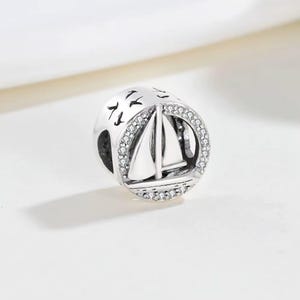 May include: A round silver charm bead featuring a sailboat design in the centre. The circular frame is accented with small clear stones. The top portion of the bead displays engraved silhouettes of birds flying against a smooth silver surface.
