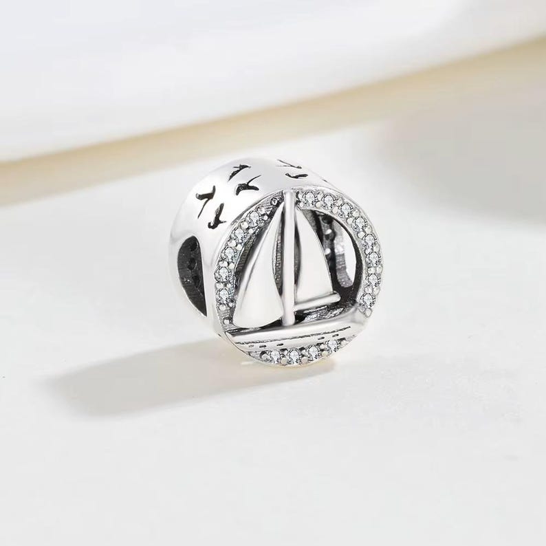 May include: A round silver charm bead featuring a sailboat design in the centre. The circular frame is accented with small clear stones. The top portion of the bead displays engraved silhouettes of birds flying against a smooth silver surface.