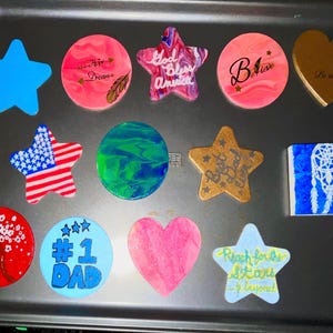 May include: A collection of twelve decorative shapes including stars, circles, hearts, and a square, featuring various designs like the American flag, a dreamcatcher, floral patterns, and text such as God Bless America, Believe, and #1 Dad.