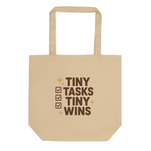 May include: Beige tote bag with the text "TINY TASKS TINY WINS" in brown. The design includes check boxes and starbursts. The bag has two handles and is made of a natural-colored fabric.