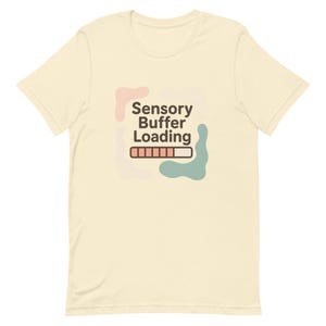 May include: Cream-colored t-shirt with the text "Sensory Buffer Loading" in black, above a loading bar graphic. The design includes abstract shapes in pink and teal. The shirt is made of a soft material.