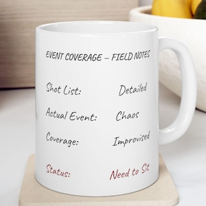 May include: White ceramic mug with the text "EVENT COVERAGE - FIELD NOTES". The text includes "Shot List: Detailed", "Actual Event: Chaos", "Coverage: Improvised", and "Status: Need to Sit". The mug sits on a beige coaster.