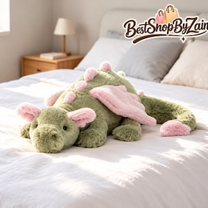 May include: A plush dragon toy with sage green fur and soft pink accents on its ears, wings, tail tip, and back spikes. The creature has dark eyes and a textured, fuzzy appearance. The text BestShopByZain is visible in the upper right corner.