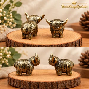 Op de afbeelding: Twee kleine, antiek goudkleurige metalen beeldjes van Highland-runderen, elk met een textuur van vacht en gebogen hoorns. De beeldjes staan op een ronde houten schijf en tonen de dieren zowel van voren als van opzij.