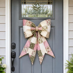 May include: A large decorative bow made of quilted fabric featuring a patchwork design. The pattern consists of alternating squares in shades of dusty pink, sage green, cream, and brown, with floral and gingham prints. The bow has two long tails.