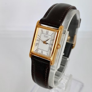 May include: A rectangular gold-tone Seiko Quartz watch with a white face and black Roman numerals. The watch has a brown leather band. The watch is displayed on a clear plastic stand.