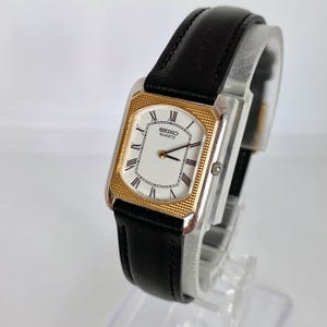 May include: A rectangular Seiko Quartz watch with a gold-tone bezel and a white face featuring black Roman numerals. The watch has a black leather band and a silver-tone case.