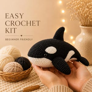 Puede incluir: Una orca de ganchillo en blanco y negro, sostenida en las manos. La imagen incluye el texto "EASY CROCHET KIT" y "BEGINNER FRIENDLY". También se ven ovillos de lana en una cesta y un ganchillo.