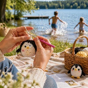May include: A person crocheting a black and white penguin toy with a pink crochet hook. A finished penguin toy sits next to a basket filled with yarn. The scene is set outdoors near a lake, with children playing in the water.