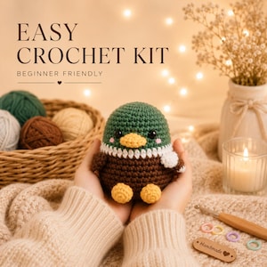 May include: A handmade crochet duckling kit held in hands, with the text "EASY CROCHET KIT BEGINNER FRIENDLY". The duckling is green, brown, and yellow, with a white collar. A basket of yarn, a candle, and crochet supplies are also visible.