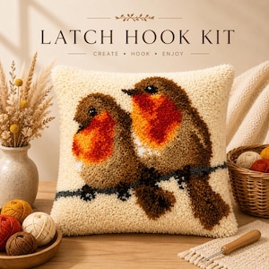 May include: A square latch hook kit pillow featuring two brown birds with orange-red chests perched on a branch against a cream background. The text LATCH HOOK KIT CREATE • HOOK • ENJOY is printed above the pillow. The design is textured and plush.