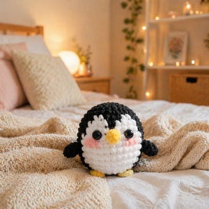 May include: A handmade, crocheted penguin with a black head, white body, and pink cheeks sits on a bed. The penguin has a yellow beak and feet, and black button eyes. The bed is covered with a cream-colored knitted blanket.