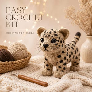May include: A crocheted leopard cub with black spots, on a cream blanket. The image includes yarn balls in a basket, a crochet hook, and text that reads "EASY CROCHET KIT" and "BEGINNER FRIENDLY".