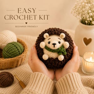 Peut inclure: Un hérisson crocheté avec un visage crème, un corps marron et une écharpe verte est tenu dans les mains. Le texte "EASY CROCHET KIT" et "BEGINNER FRIENDLY" sont affichés au-dessus. Un panier de fil et une bougie allumée sont en arrière-plan.