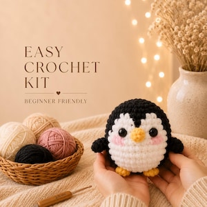 May include: A crochet kit with a handmade penguin. The penguin is black, white, and yellow with pink cheeks. The kit includes yarn in cream, pink, and black, a crochet hook, and the text "EASY CROCHET KIT BEGINNER FRIENDLY."