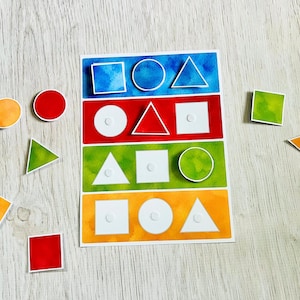 May include: A rectangular educational board featuring four horizontal rows of blue, red, green, and orange, each containing white outlines of a square, circle, and triangle. Several loose, colourful paper shapes including circles, squares, and triangles are scattered around the board.