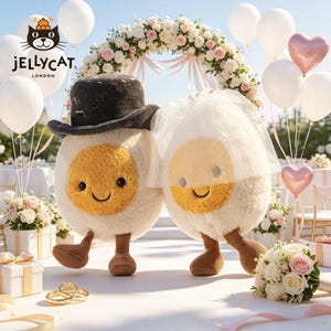 May include: Two plush egg characters, one wearing a top hat and the other a veil, stand at a wedding. The eggs have smiling faces and corduroy legs. The background includes a floral arch, balloons, and wedding decor. The Jellycat logo is visible.