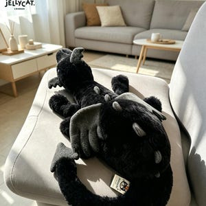 May include: A black plush dragon toy with grey wings and accents, lying on a grey chair. The dragon has a long tail and a tag from Jellycat London. The plush toy is soft and fluffy, ideal for children.