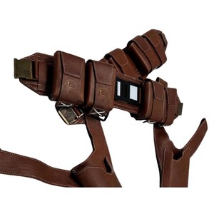 K&ouml;nnte beinhalten: Ein brauner Leder-G&uuml;rtel mit mehreren aufgesetzten Taschen, die gepr&auml;gte kreisf&ouml;rmige Logos tragen, einer zentralen schwarz-wei&szlig;en Schnalle und zwei integrierten holsterartigen Beinbefestigungen mit wei&szlig;en Ziern&auml;hten.