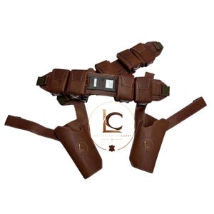 K&ouml;nnte beinhalten: Ein brauner Leder-G&uuml;rtel mit mehreren rechteckigen Taschen, einem zentralen Schnallenmechanismus und zwei angebrachten Holster-H&uuml;llen. Der Artikel verf&uuml;gt &uuml;ber wei&szlig;e N&auml;hte und ein kreisf&ouml;rmiges Logo mit dem Text LEATHERCRAFTEDCOART.