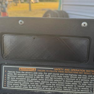 May include: A black golf cart dashboard with a warning label and safety instructions. The label includes text about operation, maximum payload of 800 lbs, and safety precautions. Keys hang from the dashboard.