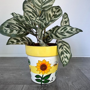 Peut inclure: Une plante en pot aux feuilles panachées vert foncé et blanc dans un pot en céramique blanc. Le pot présente un bord peint en jaune et une bande horizontale jaune, décoré d'un tournesol peint à la main, de feuilles vertes et d'une abeille.
