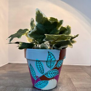 Pode incluir: Um pequeno vaso de flores de terracota pintado à mão com uma planta de hera verde e branca. O vaso é pintado com uma parte superior azul clara, uma parte média castanha e uma parte inferior rosa, com desenhos de folhas turquesas. A planta tem folhas verdes com bordas brancas.