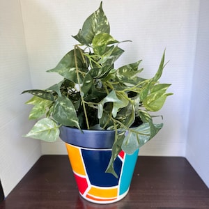 May include: A potted artificial plant with variegated green and white heart-shaped leaves. The plant is housed in a tapered ceramic pot featuring a geometric pattern of bold orange, dark blue, and bright turquoise sections with a dark blue rim.
