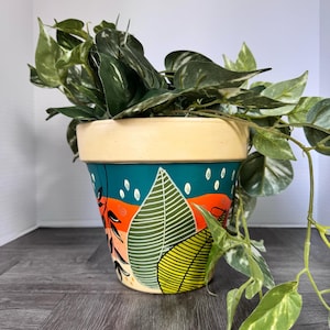 May include: A terracotta plant pot with a cream-colored rim and a hand-painted exterior featuring a teal background, orange horizontal bands, and stylized green leaf illustrations with white raindrop accents, containing a trailing green pothos plant.