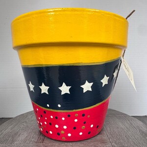 May include: A tapered plant pot featuring a wide yellow rim, a central navy blue band decorated with a row of white stars, and a red base section adorned with scattered white, black, and gold polka dots. A small paper tag hangs from the side.