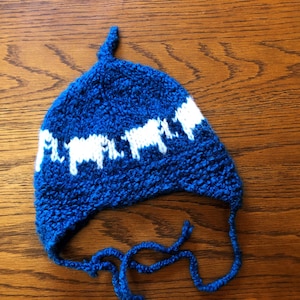 May include: A blue knitted hat with a white pattern of sheep. The hat has a long tie that can be tied under the chin.
