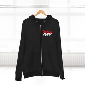 Könnte beinhalten: Schwarzer Kapuzenpullover mit Reißverschluss und dem Text "Chicago MADE" in Rot und Weiß. Der Hoodie hat einen durchgehenden Reißverschluss und eine Kapuze. Das Design befindet sich auf der oberen linken Brust. Der Hoodie hängt an einem Holzbügel.