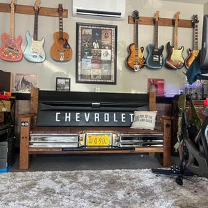May include: A rustic bench made from a Chevrolet truck tailgate, with the brand name in white lettering. The bench has a license plate and is surrounded by guitars hanging on the wall, creating a music-themed space.