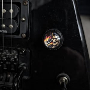 May include: Close-up of a black electric guitar with a skull-themed button. The button features a skull with a pompadour hairstyle holding a microphone, surrounded by flames. The guitar has black hardware and a toggle switch.