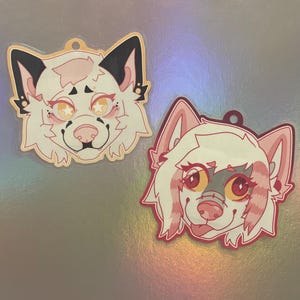 May include: Two acrylic charms featuring stylised animal head illustrations. The top charm depicts a white creature with black ears and star-shaped eyes. The bottom charm shows a white creature with pink accents, red eyes, and a small tongue sticking out.