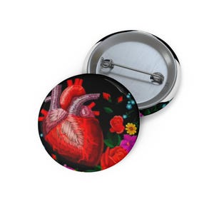 Könnte beinhalten: Ein runder Ansteckbutton mit einer detaillierten Illustration eines roten anatomischen Herzens, umgeben von leuchtend roten Rosen, kleinen gelben Blumen und violetten Blüten auf schwarzem Grund. Die Rückseite zeigt eine silberne Metallnadel.